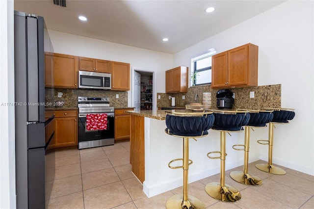 kitchen with light tile patterned floors, a peninsula, appliances with stainless steel finishes, brown cabinetry, and a kitchen bar