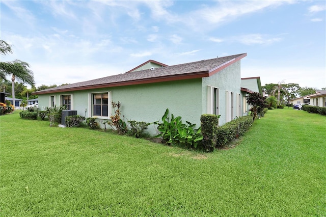 rear view of house featuring roof with shingles, a yard, central AC unit, and stucco siding