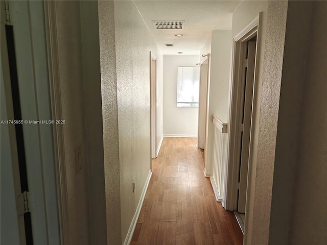 hall with a textured wall, light wood-style flooring, visible vents, and baseboards