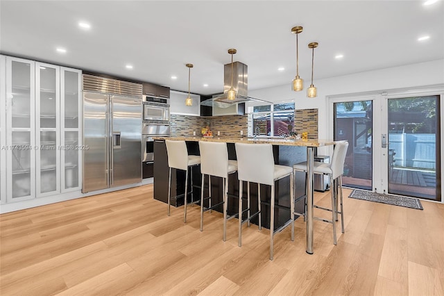 kitchen featuring island exhaust hood, light wood-style floors, appliances with stainless steel finishes, decorative backsplash, and a kitchen bar