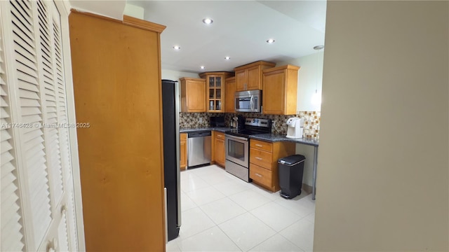 kitchen with brown cabinetry, stainless steel appliances, glass insert cabinets, dark countertops, and backsplash