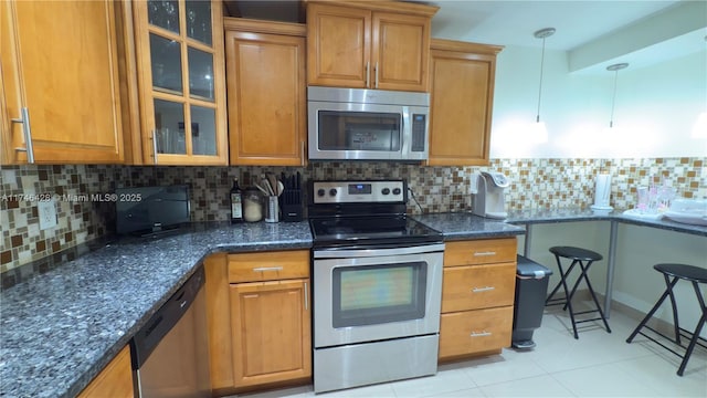 kitchen with glass insert cabinets, dark stone counters, appliances with stainless steel finishes, brown cabinetry, and decorative backsplash
