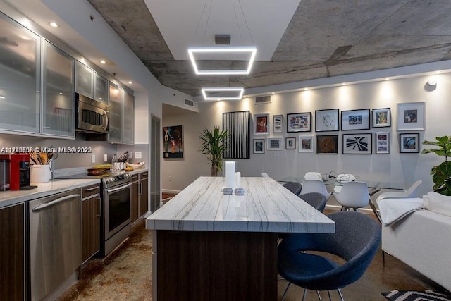 kitchen with stainless steel appliances, light countertops, glass insert cabinets, a kitchen island, and dark brown cabinets