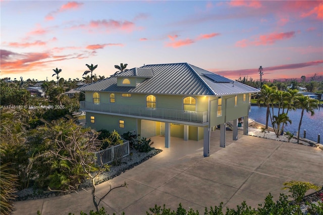 back of property at dusk with concrete driveway, a water view, roof mounted solar panels, a balcony, and a carport