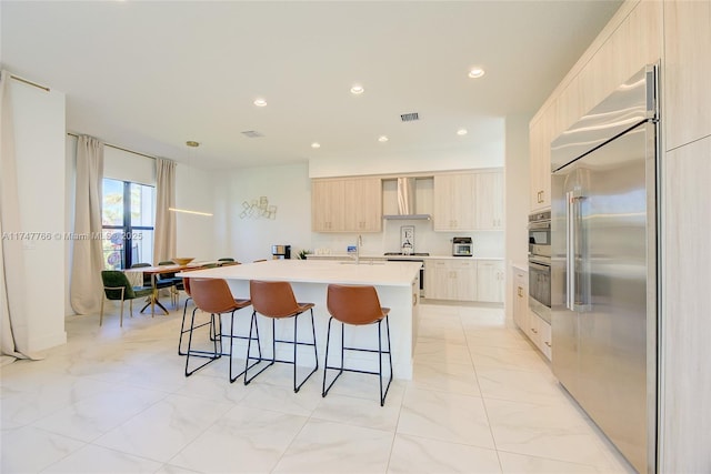 kitchen featuring a kitchen island with sink, light countertops, wall chimney range hood, light brown cabinetry, and stainless steel built in refrigerator