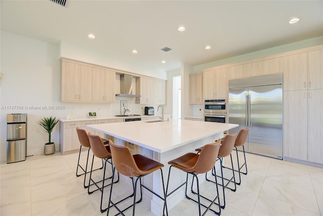 kitchen with light countertops, appliances with stainless steel finishes, a kitchen island with sink, a sink, and wall chimney exhaust hood