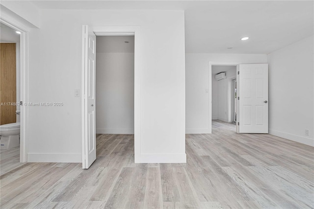 unfurnished bedroom with a walk in closet, a wall unit AC, ensuite bathroom, light wood-type flooring, and baseboards