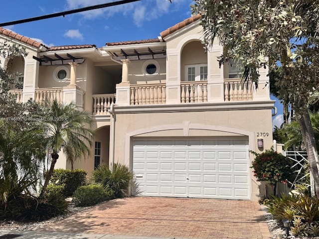 mediterranean / spanish-style home with decorative driveway, a tile roof, stucco siding, an attached garage, and a balcony