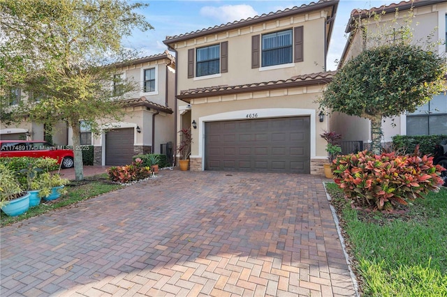 mediterranean / spanish-style house with an attached garage, a tile roof, stone siding, decorative driveway, and stucco siding