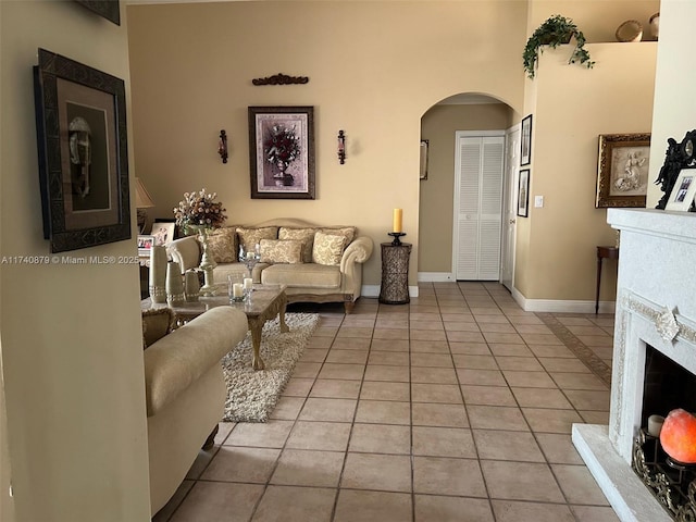 living area featuring arched walkways, a fireplace with raised hearth, light tile patterned floors, and baseboards