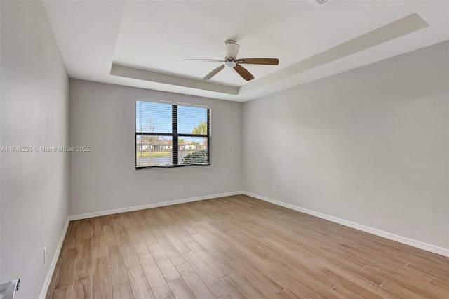 empty room featuring light wood-style flooring, a tray ceiling, ceiling fan, and baseboards