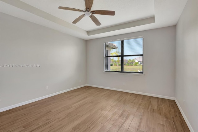 empty room with ceiling fan, a raised ceiling, light wood-style flooring, and baseboards