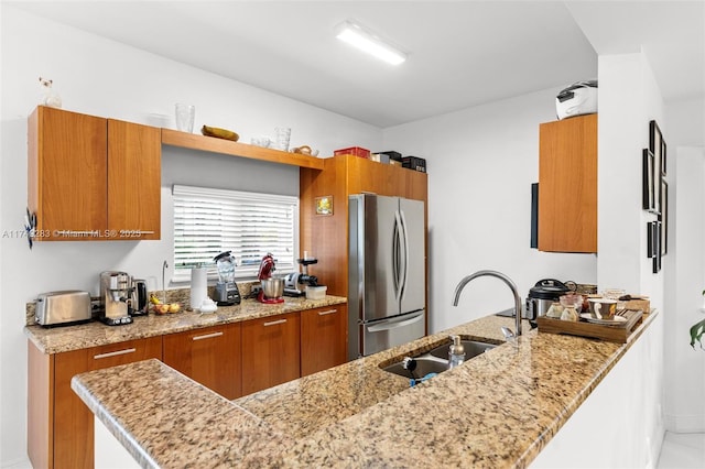 kitchen featuring a sink, a peninsula, brown cabinets, freestanding refrigerator, and light stone counters