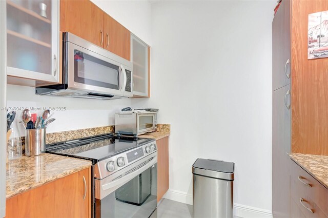 kitchen with brown cabinets, glass insert cabinets, stainless steel appliances, and light stone counters