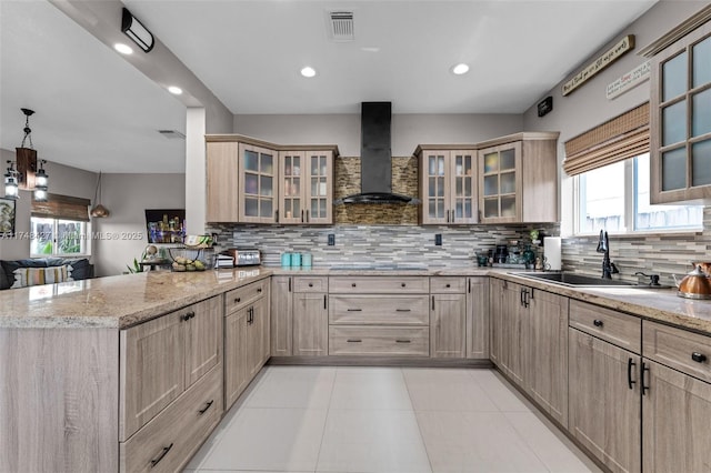 kitchen featuring wall chimney exhaust hood, glass insert cabinets, a peninsula, pendant lighting, and a sink