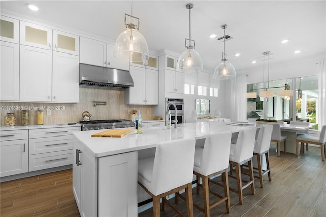 kitchen featuring under cabinet range hood, a center island with sink, glass insert cabinets, and a breakfast bar area