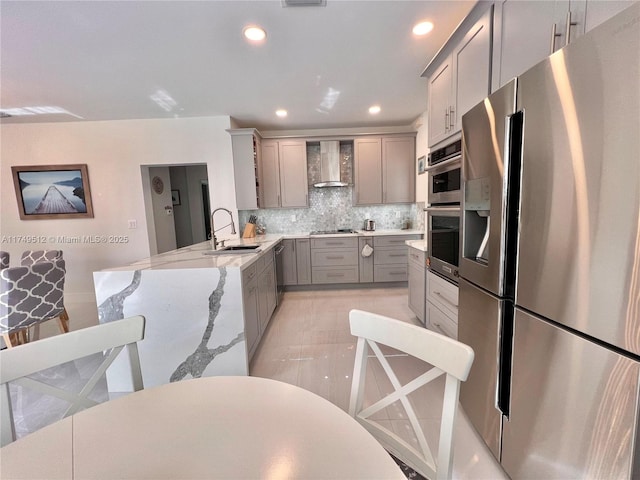 kitchen featuring wall chimney exhaust hood, appliances with stainless steel finishes, a peninsula, gray cabinets, and a sink