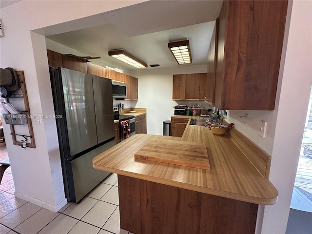 kitchen with brown cabinetry, appliances with stainless steel finishes, a peninsula, light countertops, and a sink