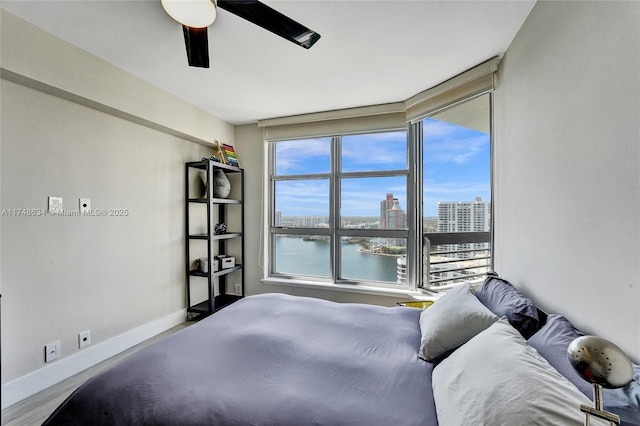 bedroom featuring baseboards, a ceiling fan, wood finished floors, a water view, and a view of city