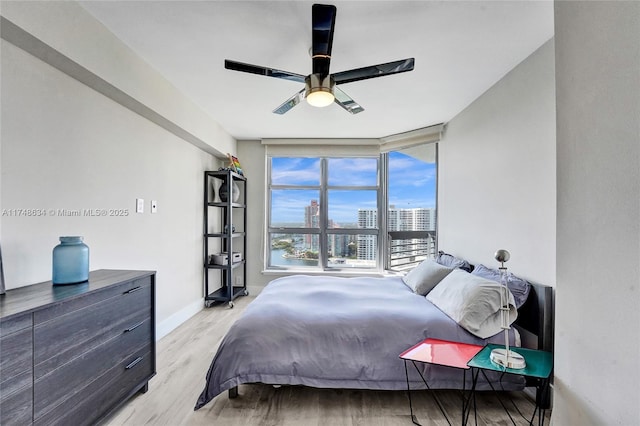 bedroom with baseboards, ceiling fan, light wood-style flooring, a view of city, and a wall of windows