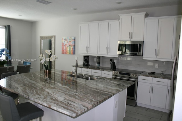 kitchen with decorative backsplash, appliances with stainless steel finishes, white cabinets, a sink, and an island with sink