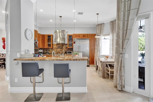 kitchen featuring glass insert cabinets, brown cabinets, hanging light fixtures, stainless steel built in refrigerator, and exhaust hood