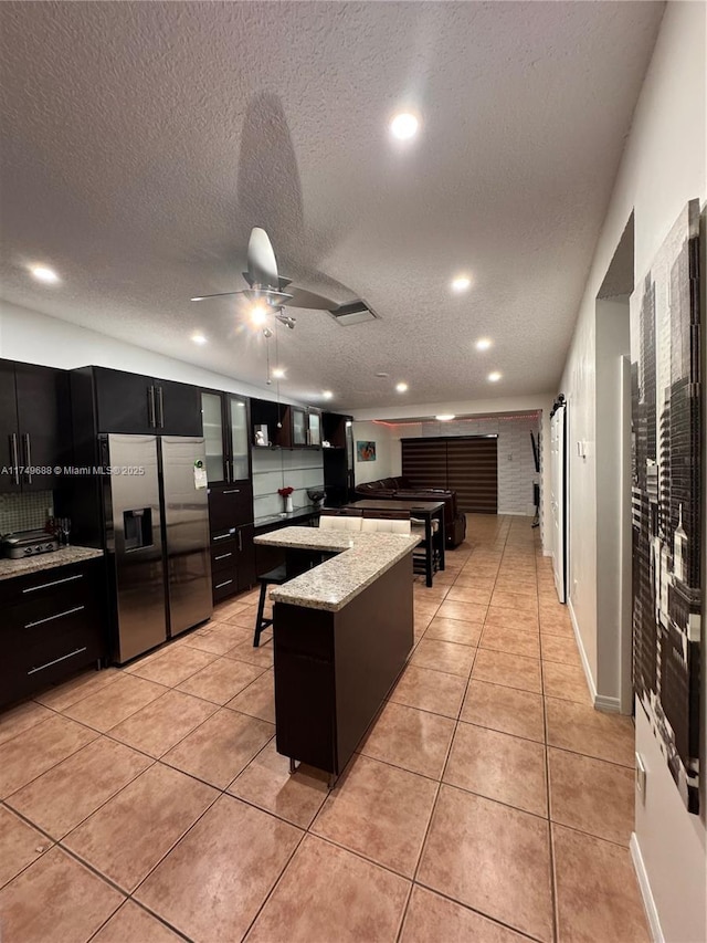 kitchen featuring a breakfast bar area, light tile patterned flooring, dark cabinetry, a center island, and stainless steel fridge