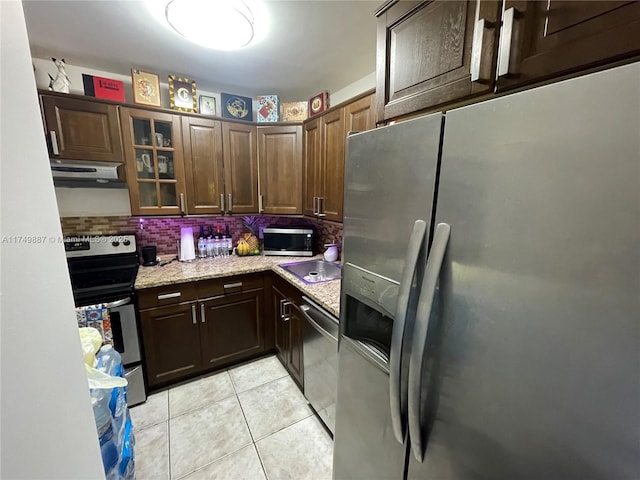 kitchen with light tile patterned floors, under cabinet range hood, stainless steel appliances, decorative backsplash, and glass insert cabinets