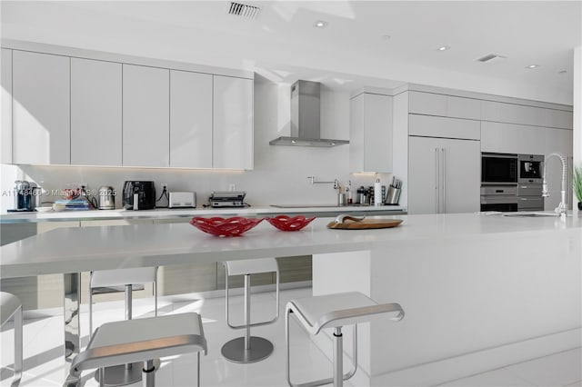 kitchen featuring built in appliances, a kitchen breakfast bar, light countertops, wall chimney range hood, and modern cabinets