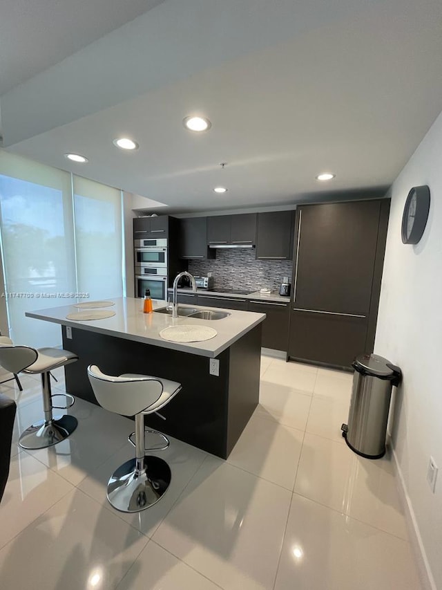 kitchen featuring decorative backsplash, a kitchen island with sink, light countertops, a kitchen bar, and a sink