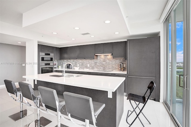 kitchen with a breakfast bar area, stainless steel double oven, a sink, light countertops, and tasteful backsplash