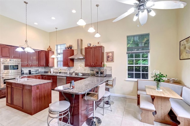 kitchen with a breakfast bar area, stainless steel appliances, hanging light fixtures, decorative backsplash, and wall chimney exhaust hood