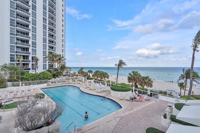 community pool with a patio area, a water view, fence, and a beach view