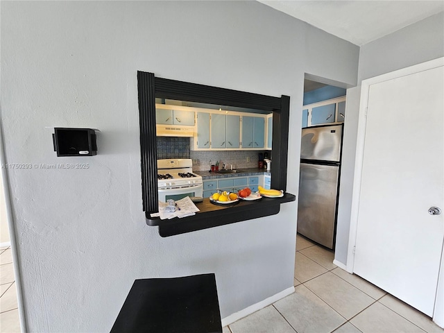 kitchen featuring light tile patterned floors, under cabinet range hood, white gas range oven, freestanding refrigerator, and decorative backsplash