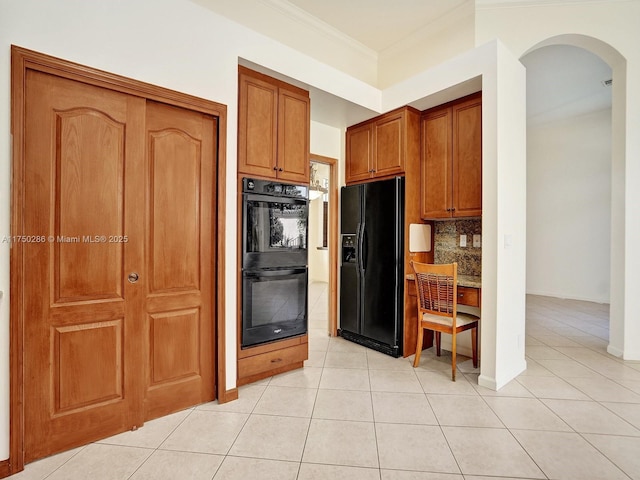 kitchen with light tile patterned floors, arched walkways, brown cabinetry, black appliances, and backsplash