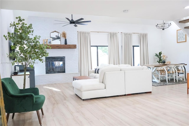 living room featuring a fireplace, light wood-style flooring, brick wall, and ceiling fan with notable chandelier