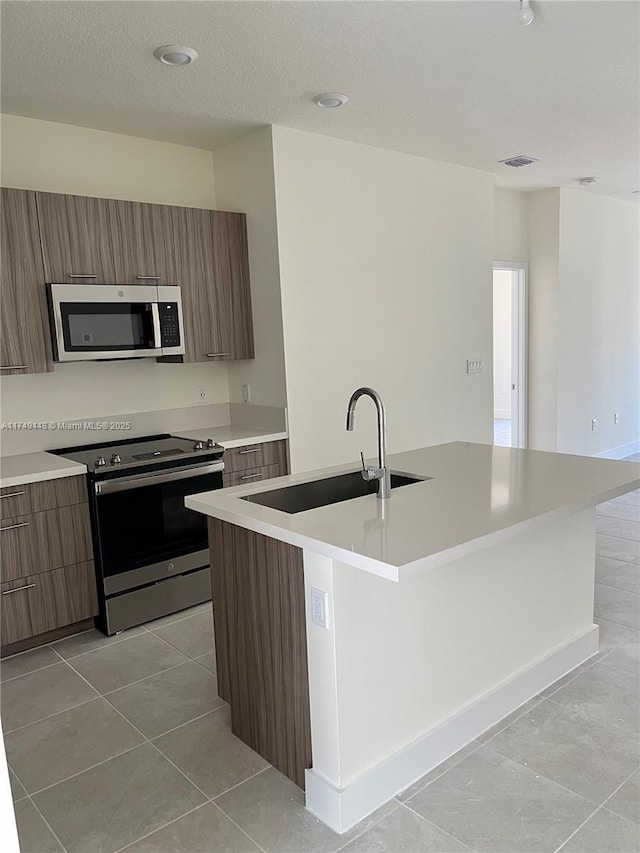 kitchen with brown cabinetry, an island with sink, appliances with stainless steel finishes, light countertops, and a textured ceiling