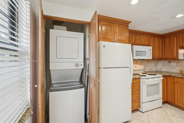 kitchen with white appliances, light tile patterned floors, brown cabinetry, light stone counters, and stacked washing maching and dryer