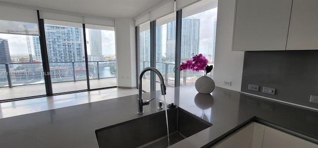 kitchen with dark countertops, white cabinetry, a wall of windows, a view of city, and a sink