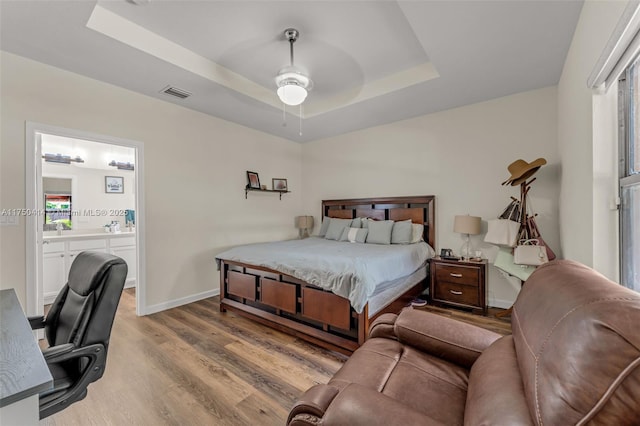 bedroom with a raised ceiling, visible vents, ensuite bathroom, light wood-style floors, and baseboards