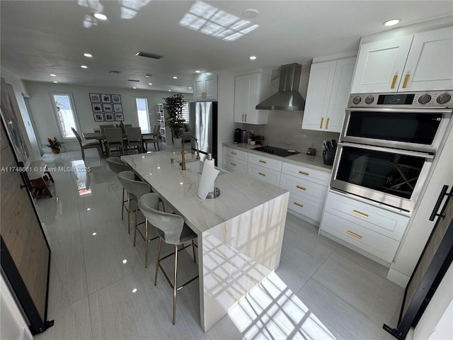 kitchen featuring wall chimney range hood, appliances with stainless steel finishes, a kitchen island with sink, and white cabinets