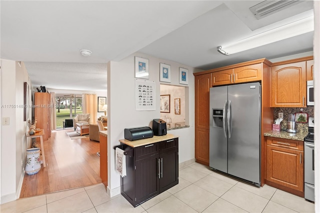 kitchen with light tile patterned floors, backsplash, appliances with stainless steel finishes, brown cabinetry, and light stone countertops