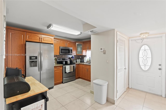 kitchen featuring light tile patterned floors, light stone counters, stainless steel appliances, brown cabinets, and glass insert cabinets