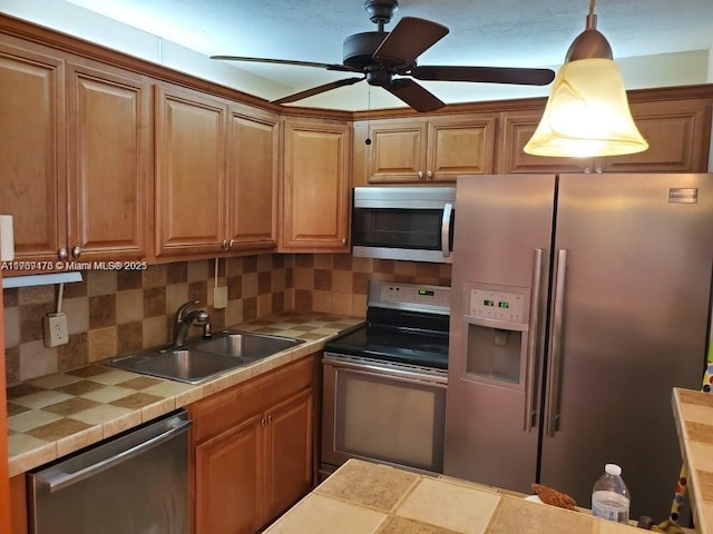 kitchen featuring tile counters, stainless steel appliances, hanging light fixtures, decorative backsplash, and a sink