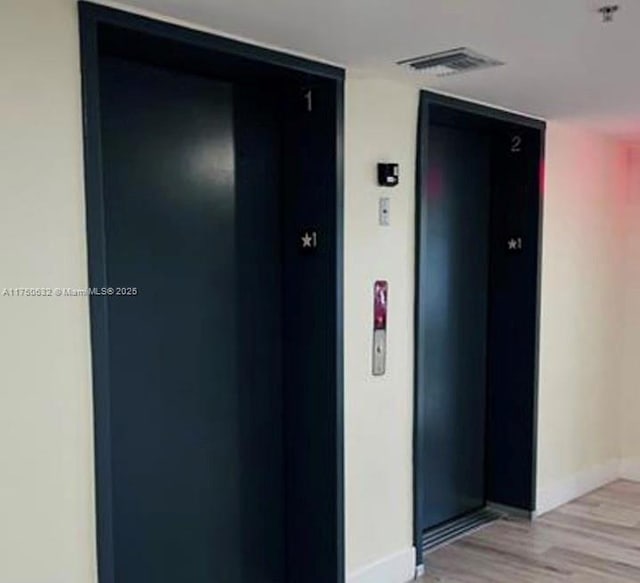 hall featuring light wood-type flooring, elevator, baseboards, and visible vents