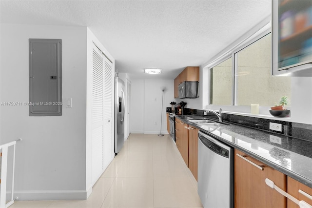 kitchen featuring stainless steel appliances, a sink, electric panel, brown cabinetry, and dark stone countertops