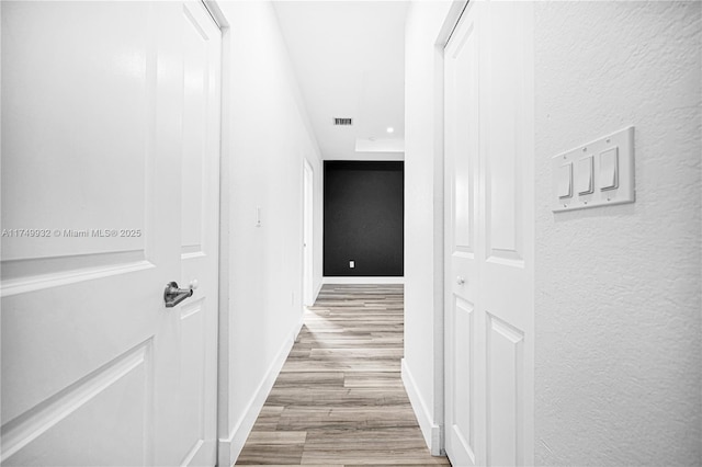 hall featuring a textured wall, light wood-type flooring, visible vents, and baseboards