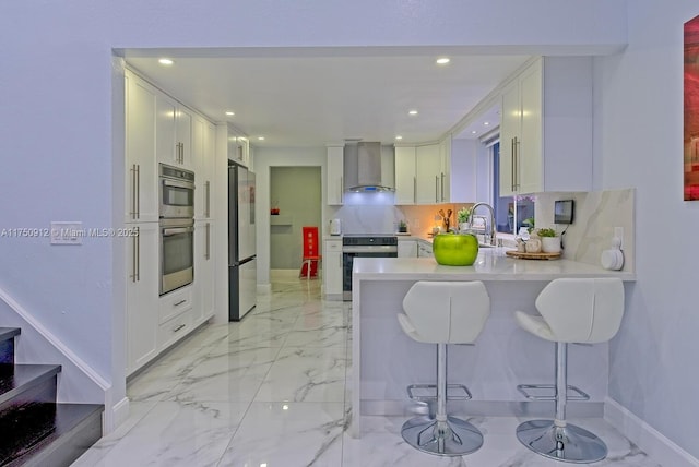 kitchen featuring a breakfast bar, white cabinets, light countertops, wall chimney range hood, and appliances with stainless steel finishes