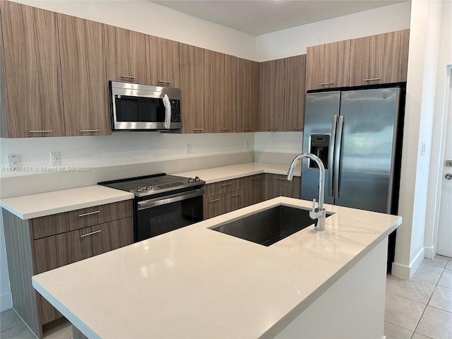 kitchen with a sink, light countertops, appliances with stainless steel finishes, brown cabinets, and an island with sink