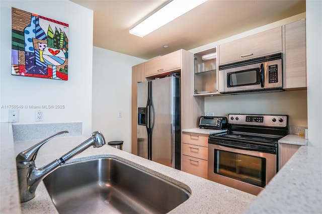 kitchen featuring appliances with stainless steel finishes, glass insert cabinets, light brown cabinets, a sink, and light stone countertops